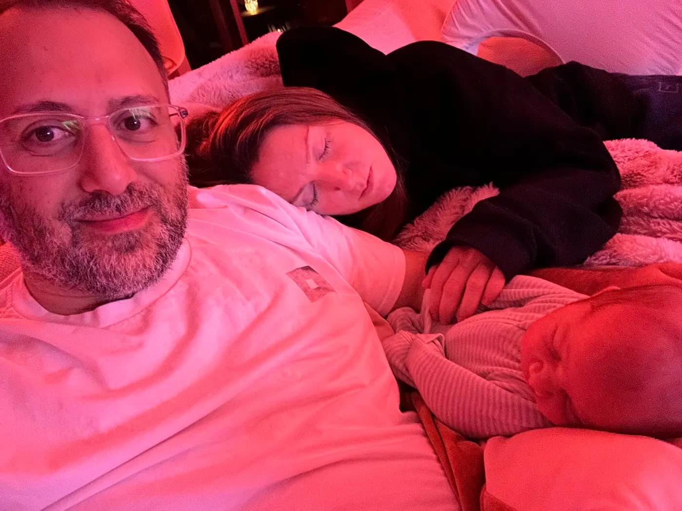 The three of us on a bed under a red night light: Kilian awake and looking at the camera, Penelope asleep with a hand resting on the baby, and the baby asleep beside her.