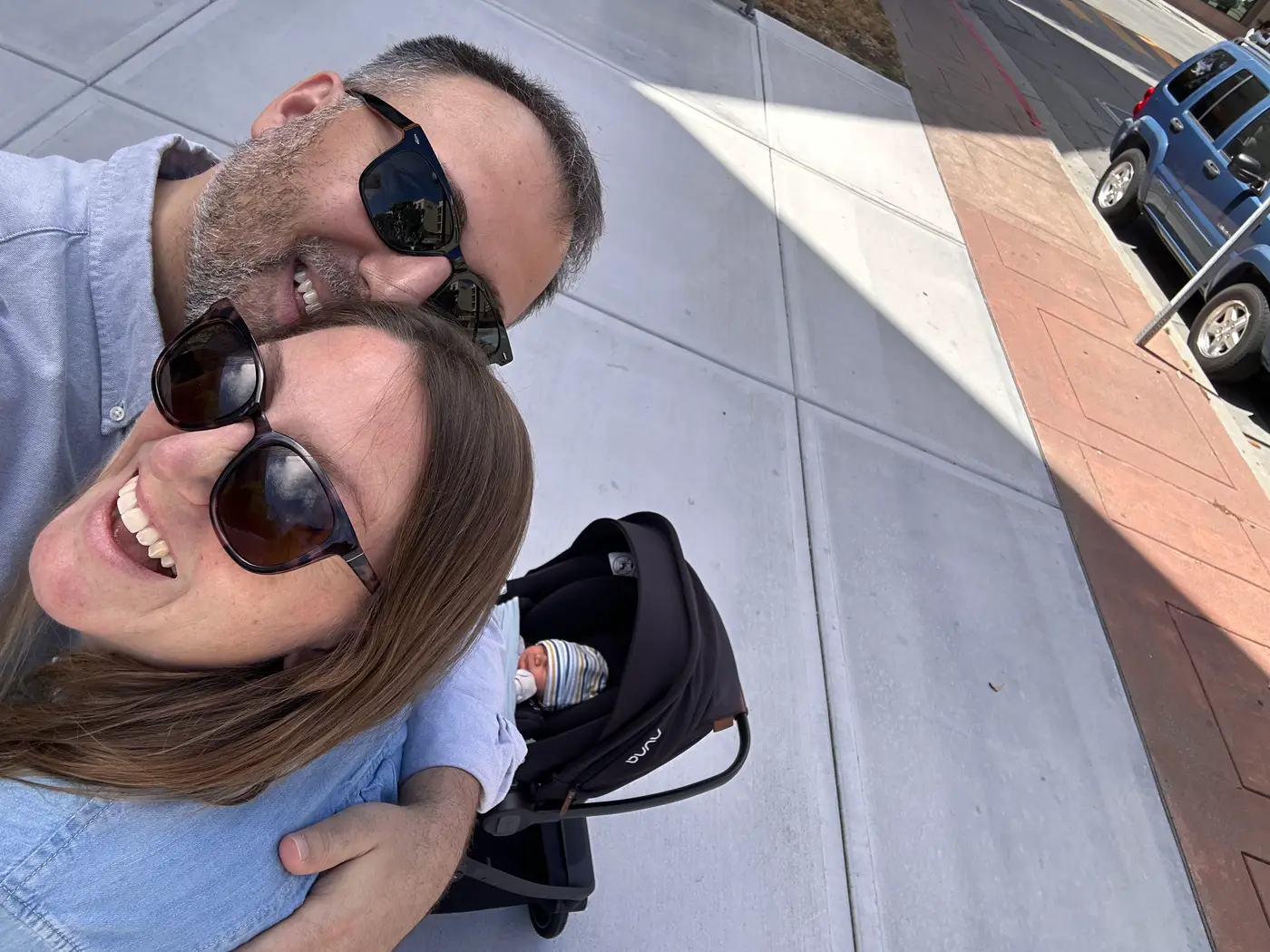 The three of us outside on a sunny pavement: Kilian and Penelope in sunglasses, laughing, with the baby asleep in a stroller beside them.